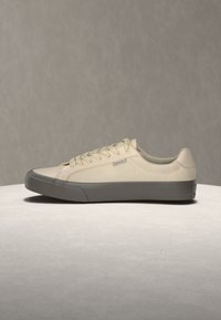 Μπεζ sneaker χαμηλού πάτου με επάνω μέρος από καμβά και χοντρή γκρι λαστιχένια σόλα, εμφανίζεται σε ουδέτερη επιφάνεια με γκρι φόντο.