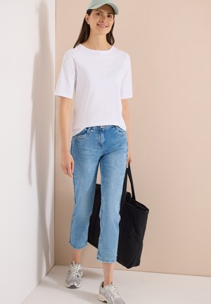 Femme portant un t-shirt blanc à manches courtes, jean court bleu clair, baskets argentées, casquette verte, tenant un sac cabas noir, debout contre un mur beige.