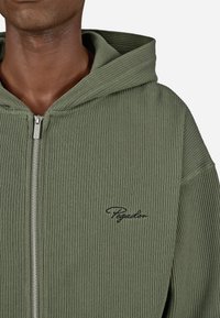 Zelená rebrovaná mikina na zips vyrobená z textúrovanej látky s veľkou kapucňou, s čiernym vyšitým logom na hrudi a strieborným zipsom.