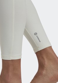 Bílé, žebrované legíny s přiléhavým designem. Disponují logem poblíž lemu. Tkanina vypadá hladce a má pružnou texturu.
