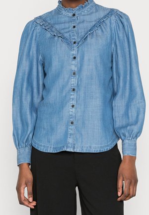 Blouse bleu clair boutonnée avec col montant à volants et empiècement, manches longues bouffantes, portée avec un pantalon noir.