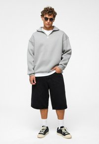 Lichtgrijze ritschool met hoge kraag, gecombineerd met losse zwarte shorts, witte sokken en zwarte sneakers met lichte accenten.