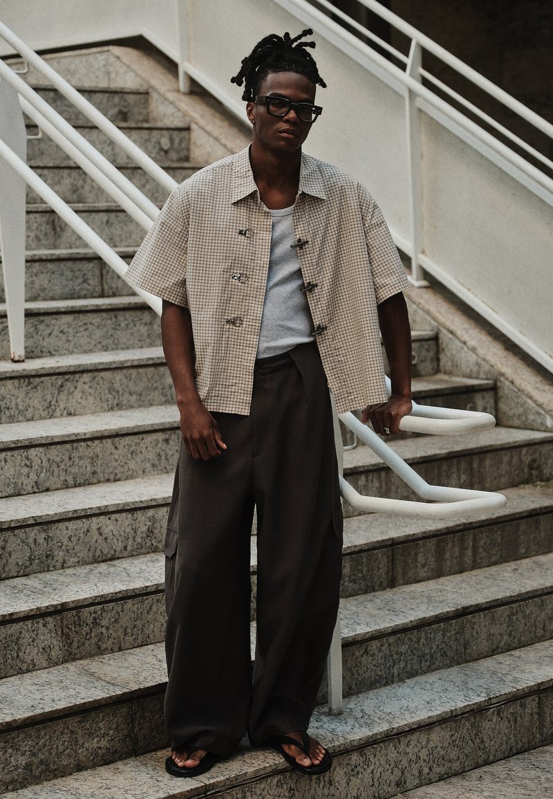 Homme avec des dreadlocks et des lunettes sombres, debout sur des escaliers en pierre, portant une chemise à manches courtes à carreaux, un débardeur gris, un pantalon large foncé et des sandales noires.