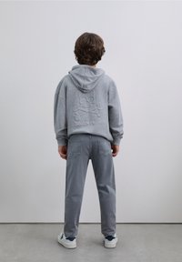 Sweat à capuche gris avec un graphisme de crâne texturé et le texte "Scalpers" au dos, associé à un pantalon gris et des baskets blanches.