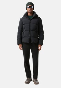 Czarna kurtka puchowa z wysokim kołnierzem, kieszeniami na zamek i kapturem. Połączona z ciemnymi spodniami, okularami przeciwsłonecznymi i zieloną czapką beanie.