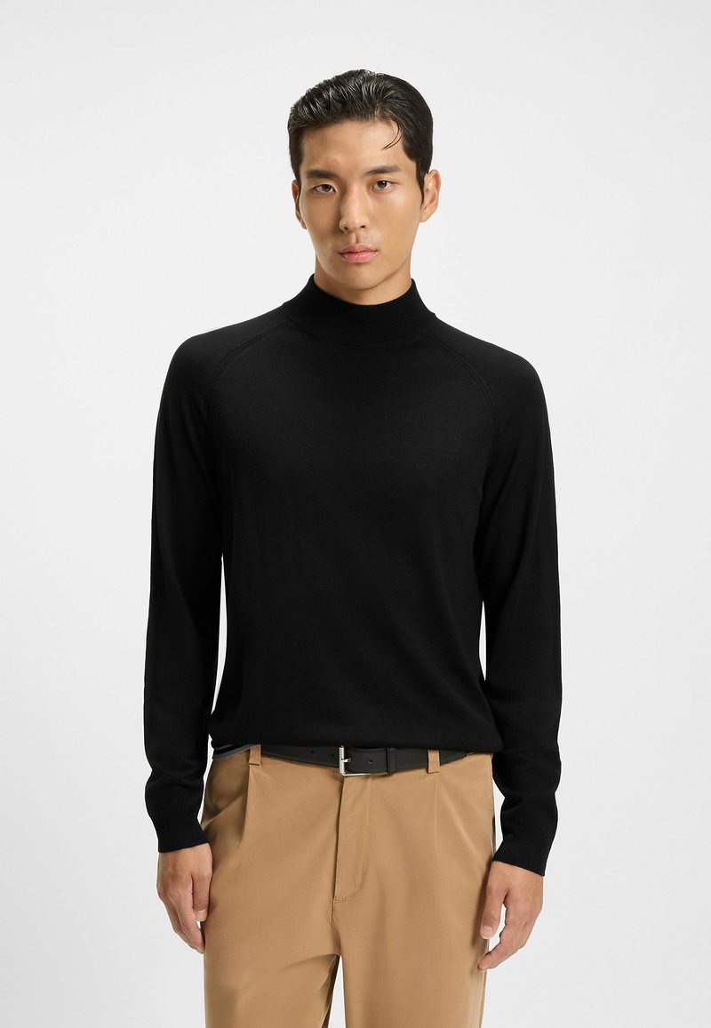 Maglione nero a collo alto in un materiale morbido, con maniche lunghe e un design aderente. Abbinato a pantaloni beige e una cintura nera.
