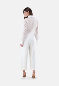 Blusa bianca con maniche lunghe trasparenti e ricamate, abbinata a pantaloni bianchi a palazzo e sandali con cinturino color beige, vista da dietro.