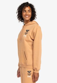 Beige Kapuzenpullover mit einer vorderen Tasche, ausgestattet mit schwarz-weißen Grafiken auf der Brust. Aus Baumwollmaterial mit einer weichen Textur.
