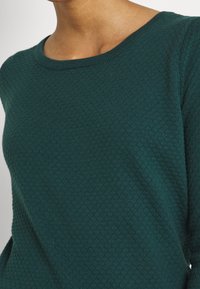 Nahaufnahme einer Person, die einen dunkelgrünen strukturierten Strickpullover mit rundem Halsausschnitt trägt.