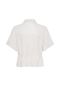 Chemise blanche à manches courtes, boutonnée, col à revers et taille cintrée, avec une broderie discrète en œillets carrés sur le tissu.