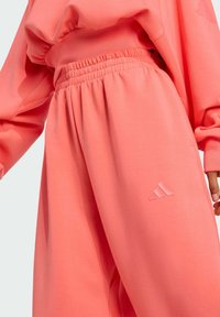 Korálovo ružová mikina a zodpovedajúce nohavice, ktoré majú množstvo zberovaných pásov, voľný strih a textúrový akcent loga na nohe.