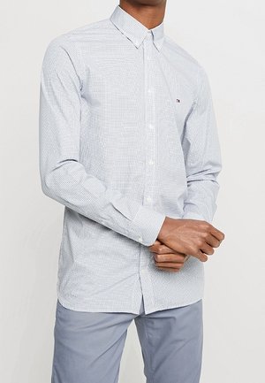 Homme portant une chemise à manches longues claire à motifs avec un petit logo sur la poitrine, associée à un pantalon gris clair.
