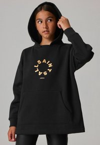 Sudadera negra de gran tamaño hecha de una tela suave, con un bolsillo frontal y un logo circular dorado que lleva el texto "SAINT". Capucha con cordones ajustables.