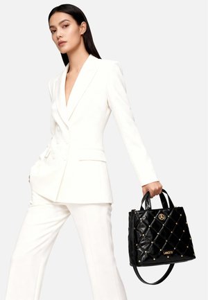 Zwarte gewatteerde handtas met gouden accenten en handvaten, met een glanzende textuur. Het accessoire wordt vastgehouden door een persoon in een wit pak.