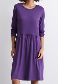 Purpurové šaty z měkkého materiálu, s dlouhými rukávy, kulatým výstřihem a staženým pasem, který přechází do délky po kolena.