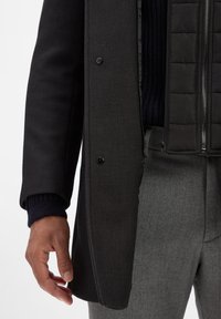 Schwarzer Mantel mit strukturiertem Stoff und einer taillierten Innenweste mit Reißverschluss, gerippten Bündchen und grauer Hose. Fokus auf Materialien und Konstruktion.