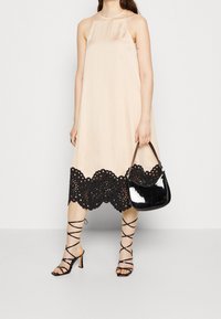 Robe sans manches beige avec un ourlet en dentelle noire à bord festonné. Assortie à des talons noirs à lanières et un sac à main noir brillant. Texture de tissu lisse.