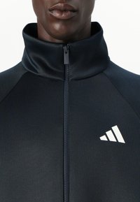 Casaco atlético preto com colarinho alto e fecho éclair frontal; apresenta um logo branco da Adidas, feito de um tecido liso e texturizado.