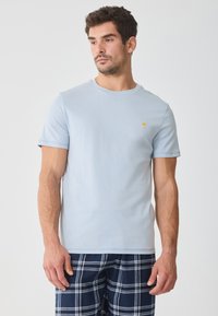 Light blue cotton t-shirt with short sleeves, round neck, and a small yellow logo on the chest, paired with navy plaid pajama bottoms.