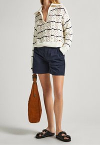 Donna che indossa un maglione bianco a maglia con strisce nere, pantaloni corti blu navy, sandali neri e tiene una borsa in suede marrone al suo fianco.