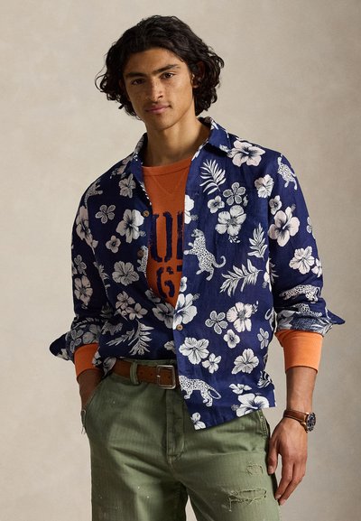 Jeune homme portant une chemise bleu marine à imprimé floral et léopard sur un sweat-shirt orange, un pantalon vert avec une ceinture marron et une montre-bracelet.