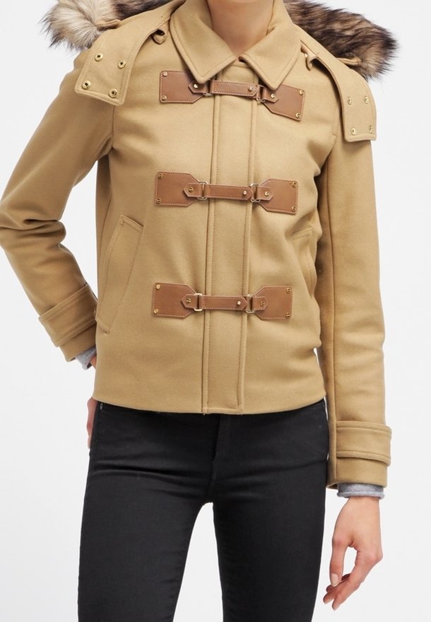 Beige jack met drie bruine leren gespsluitingen, een met bont afgewerkte capuchon en voorzakken, gedragen met zwarte broek.