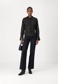 Camicia a righe bianco e nero con bottoni e maniche lunghe, abbinata a pantaloni a gamba larga in denim scuro e stivaletti neri con tacco, mentre tiene in mano una piccola borsa.