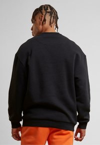 Sweatshirt en coton noir avec un col rond, des épaules tombantes et des poignets côtelés, associé à un pantalon orange. Design simple et épuré.