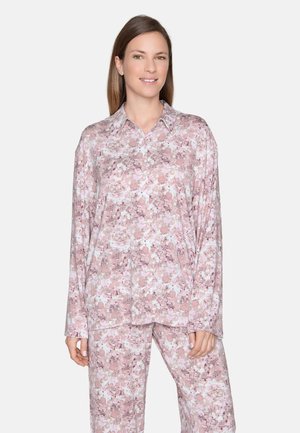 Vrouw met stijlbruin haar draagt een lichtroze pyjama met bloemenprint en lange mouwen, glimlachend en staand tegen een effen witte achtergrond.