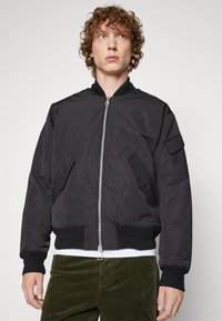 Giubbotto bomber nero in nylon con polsini e fondo a costine, chiusura con zip, tasche laterali e un piccolo logo sul petto. Il modello indossa pantaloni in velluto a coste.