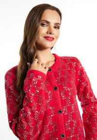 Cardigan roșu tricotat cu un model în carouri din paiete, având guler rotund, mâneci lungi și nasturi negri.