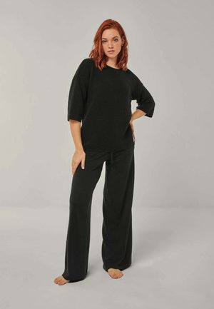 Woman with red hair stands barefoot wearing loose black pants and a black oversized ribbed sweater against a plain gray background.