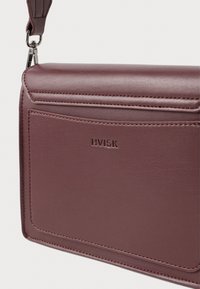 Kompaktowa torba crossbody z ciemno-brązowej skóry, z przednią kieszenią, widocznym szwem, wytłoczonym logo "HVISK" oraz odczepianym klipsem na pasek.