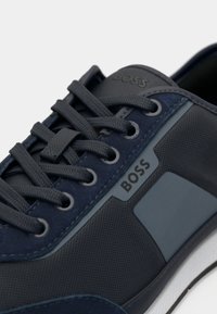 Tumsas zilas teniskurpes ar teksturētu auduma augšdaļu, gludām detaļām, kontrastējošu pelēku logotipu un saskaņotām aukliņām uz baltas zoles.