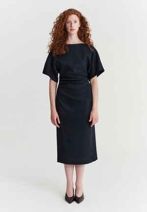 Femme aux longs cheveux roux bouclés, portant une robe midi noire et des talons pointus noirs, debout devant un fond blanc uni.