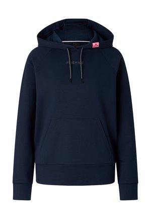 Blondynka w granatowej bluzie z kapturem i spodniach dresowych, stojąca z rękami w kieszeniach, nosząca białe masywne trampki na jednolitym tle.