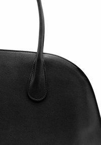 Gros plan sur un sac à main en cuir noir montrant la surface texturée et l'attache de la poignée cousue, sur fond blanc.