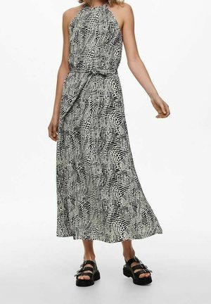 Femme portant une robe longue sans manches à motif noir et blanc avec col halter et des sandales à plateforme noires sur fond blanc.