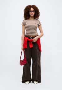 Γυναίκα με σγουρά μαλλιά που φοράει γυαλιά, καφέ ριγέ μπλουζάκι, φαρδιά σκούρα παντελόνια, κόκκινο πουλόβερ δεμένο στη μέση, κρατά κόκκινη τσάντα.