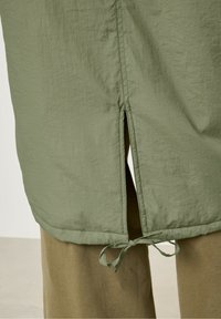 Jasnozielona, teksturowana tkanina z bocznym rozcięciem i detalem w postaci wiązania u dołu. Element jest nałożony na beżowy, miękki materiał.