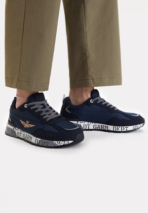 Sneakers blu navy con lacci bianchi decorati e suola intermedia bianca stampata, indossate con pantaloni beige cropped su sfondo chiaro.