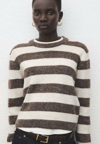 Suéter de punto a rayas marrón y crema con escote redondeado y mangas largas, llevado sobre una camisa blanca, combinado con un cinturón oscuro.