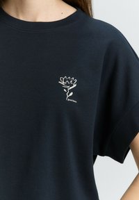 T-shirt en coton bleu marine avec un col rond et des manches courtes. Présente un graphique de fleur blanche et le texte "bonnes temps" en dessous.