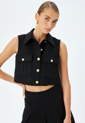 Zwarte cropped vest gemaakt van textiel met een kraag, twee voorzakken en gouden knopen. Gecombineerd met huidkleurige onderkleding zichtbaar.