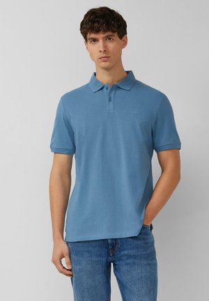 Młody mężczyzna ubrany w niebieską koszulkę polo z krótkim rękawem i niebieskie dżinsy, stojący z jedną ręką w kieszeni na jednolitym szarym tle.