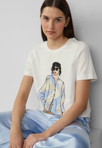 T-shirt en coton blanc avec une illustration colorée d'une femme portant des lunettes de soleil, associée à un pantalon satiné bleu clair. Manches courtes, col rond.