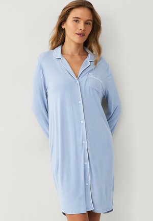 Vrouw draagt een lichtblauw nachthemd met knopen, witte biezen en een borstzak, staand tegen een eenvoudige lichtgekleurde muur.