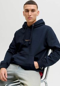 Tumesinine kapuutsiga särk, millel on känguru tasku, reguleeritav nöör ja tagasihoidlik logo tikand. Kombineeritud heledate teksadega. Tekstureeritud kangas.