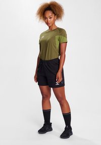 Olivengrøn atletisk T-shirt med kontrasterende mørkere grøn sidepanel, parret med sorte shorts og matchende sorte sportssokker og sko.