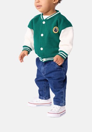 Grønn og hvit varsityjakke med striper, med en rund lapp, kombinert med blå denimbukser og hvite sneakers med røde detaljer.